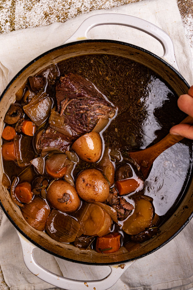 Cooked chuck roast, potatoes, carrots, onions, and mushrooms all in a broth, in a dutch oven. A hand is holding a wooden spoon, which is stirring the pot roast.