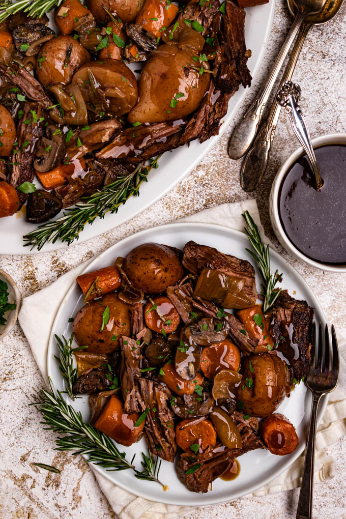 Perfect pot roast is on a white plate with gravy drizzled on top. Parsley is sprinkled over the pot roast. There are rosemary sprigs alongside the plate. A white napkin is underneath the plate, and a fork is alongside the plate. There is a platter of pot roast in the upper left hand corner of the photo, along with serving spoons to the right. A bowl of gravy with a spoon in it is to the right.