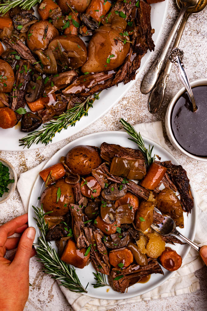 A plate of perfect pot roast with gravy drizzled on top. The plate is garnished with rosemary sprigs, and chopped parsley leaves. There are hands holding the plate, and a fork stabbing a potato. There is a platter of the pot roast in the upper left corner of the photo. To the right is a bowl of the gravy with a spoon.