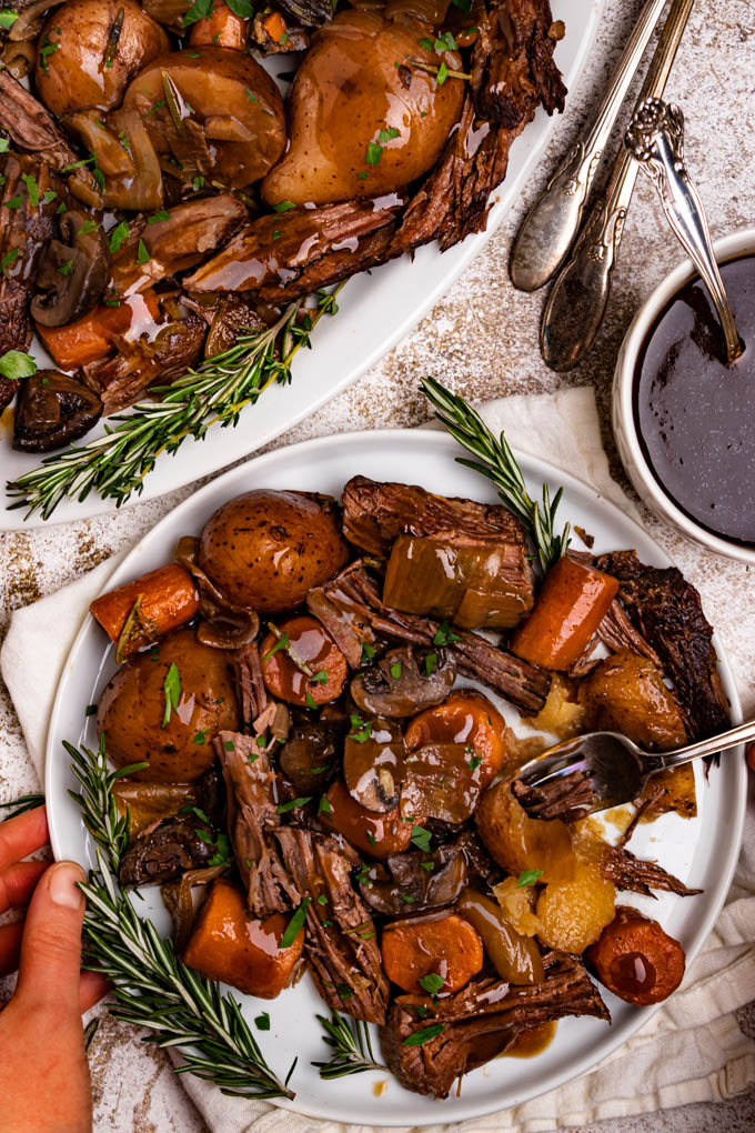 A closeup shot of perfect pot roast on a plate. A hand is holding the plate, and a fork is stabbing a potato. A platter of pot roast is in the upper left hand corner, along with serving utensils. A bowl of gravy is off to the right.