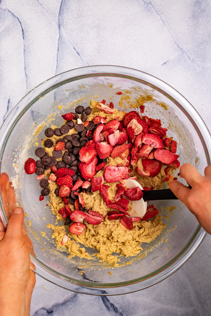 Cookie dough in a mixing bowl with freeze dried strawberries and chocolate chips being stirred in by a spatula - hands are holding the bowl.