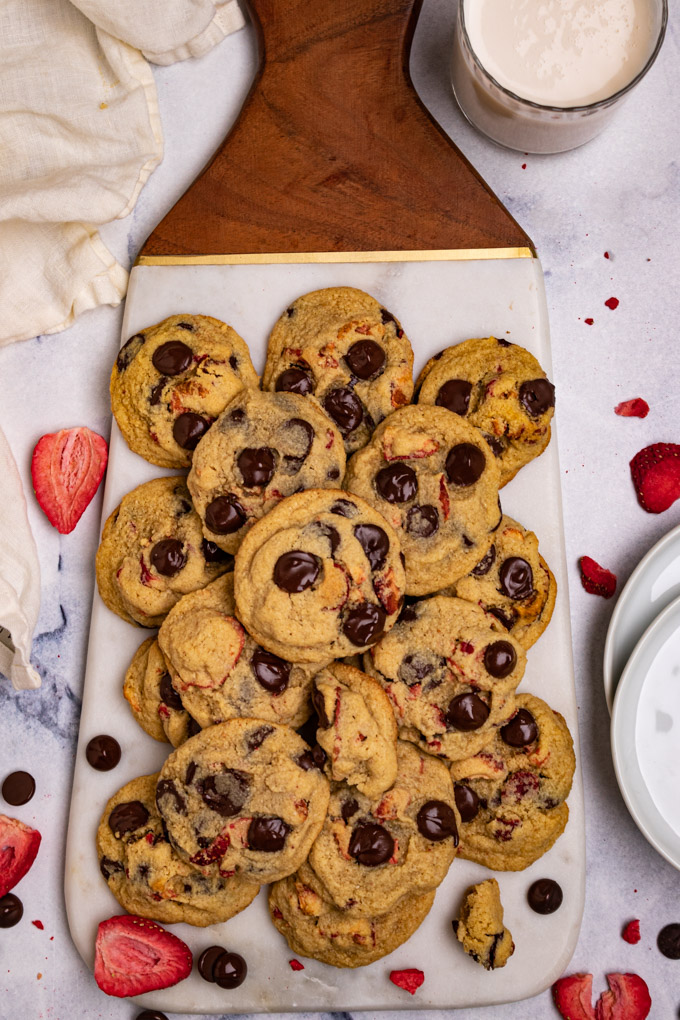 Overhead shot of strawberry chocolate chip cookies on a marble tray. A glass of milk is in the corner. Plates are off to the side. Freeze dried strawberries, and chocolate chips are scattered around.