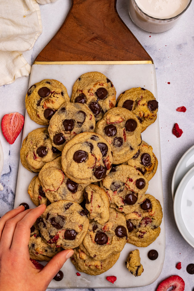 Overhead shot of strawberry chocolate chip cookies on a marble tray. A hand is picking up one of the cookies. 