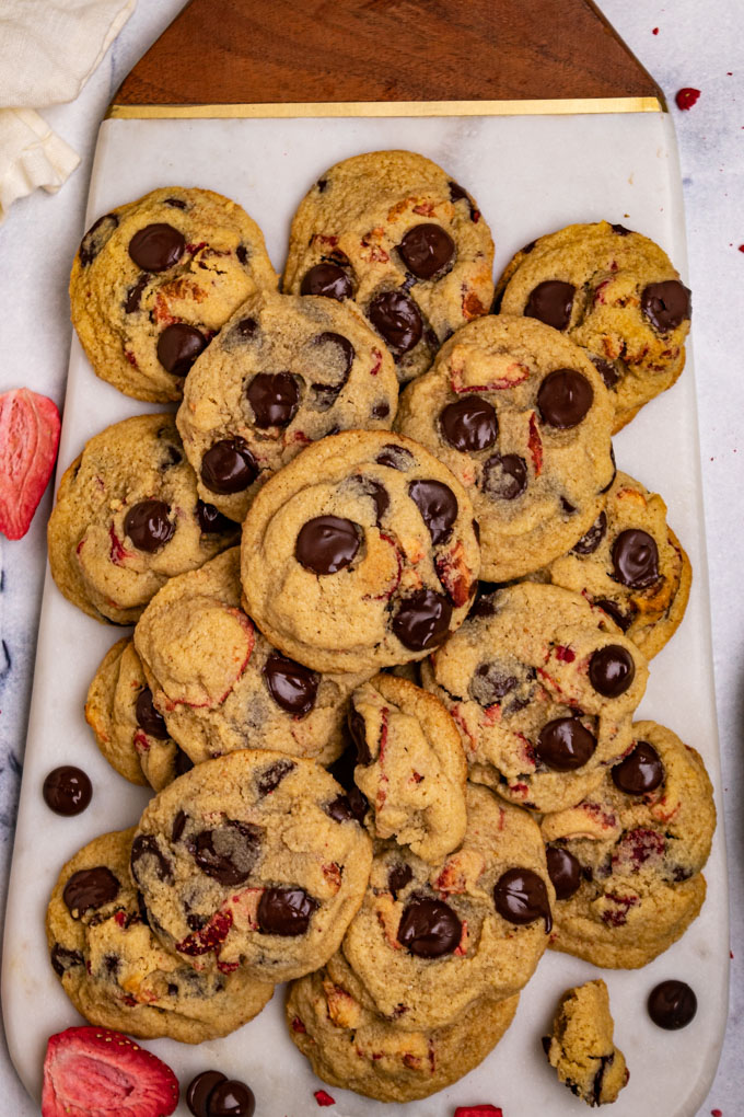 strawberry chocolate chop cookies are layered on a marble cutting board. A few freeze dried strawberries, and chocolate chips are scattered around.