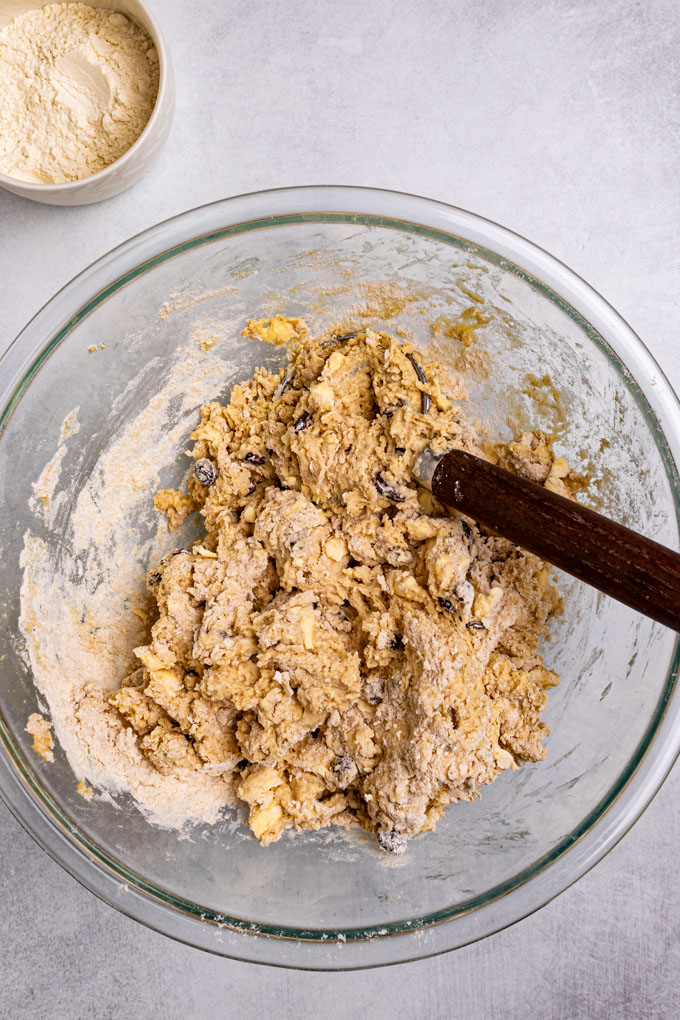 A mixing bowl of a shaggy dough mixed together. A small bowl of flour is off to the side.