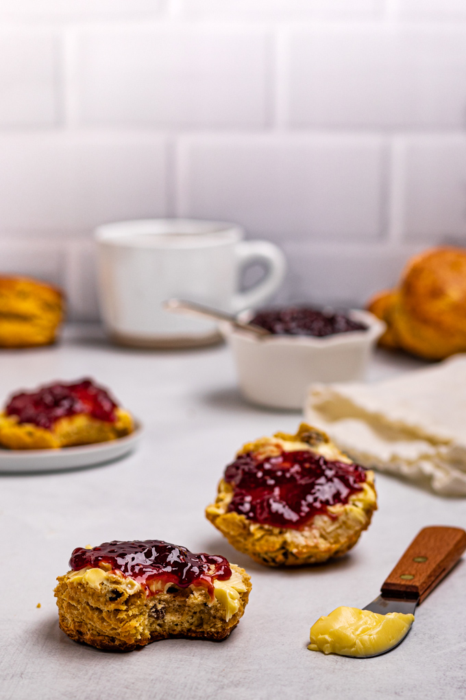 Straight on shot of Irish scones. One scone is split in half and has butter and jam spread on it, it has a bite taken out of it. A mug of tea is in the background along with more scones.