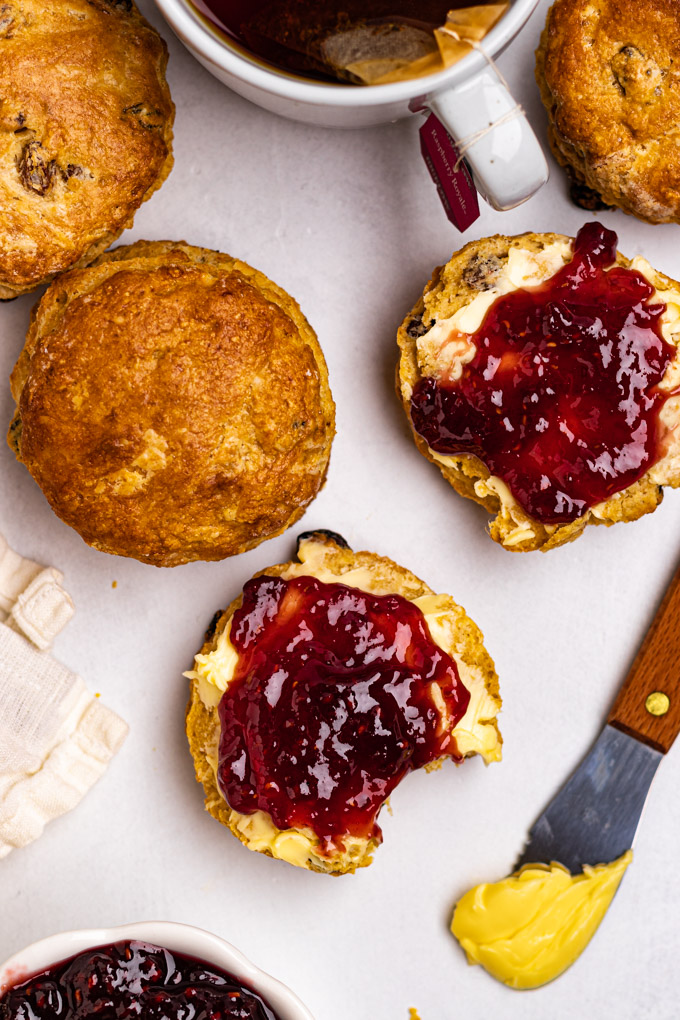 Overhead photo of Irish scones. One is split in half and has butter and jam spread on it, with a bite taken out of it. More scones are scattered around that scone. A spreader has butter on it. There is a mug of tea.