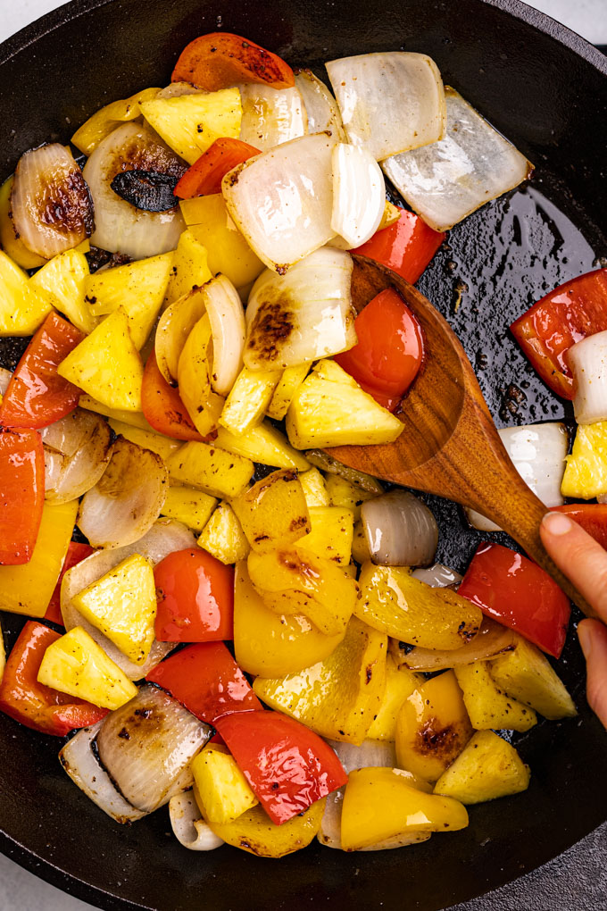 Onions, bell peppers, and pineapple in a skillet being stirred by a wooden spoon.