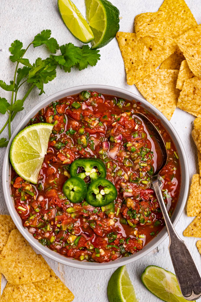 Homemade salsa in a bowl with a spoon in it. There are sliced jalapenos in the center, and a lime off to the side. Tortilla chips, lime slices, and cilantro are scattered in the background.