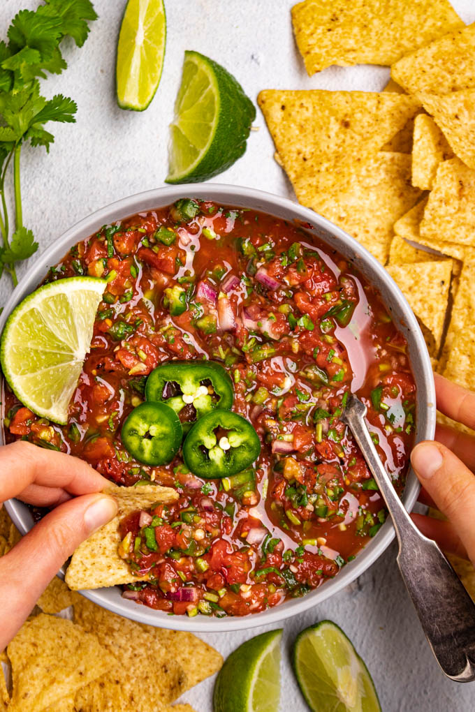 A bowl of homemade salsa with slices of jalapeno on top, along with a lime. A hand is dipping a chip into the salsa. Chips and limes are scattered throughout the background.