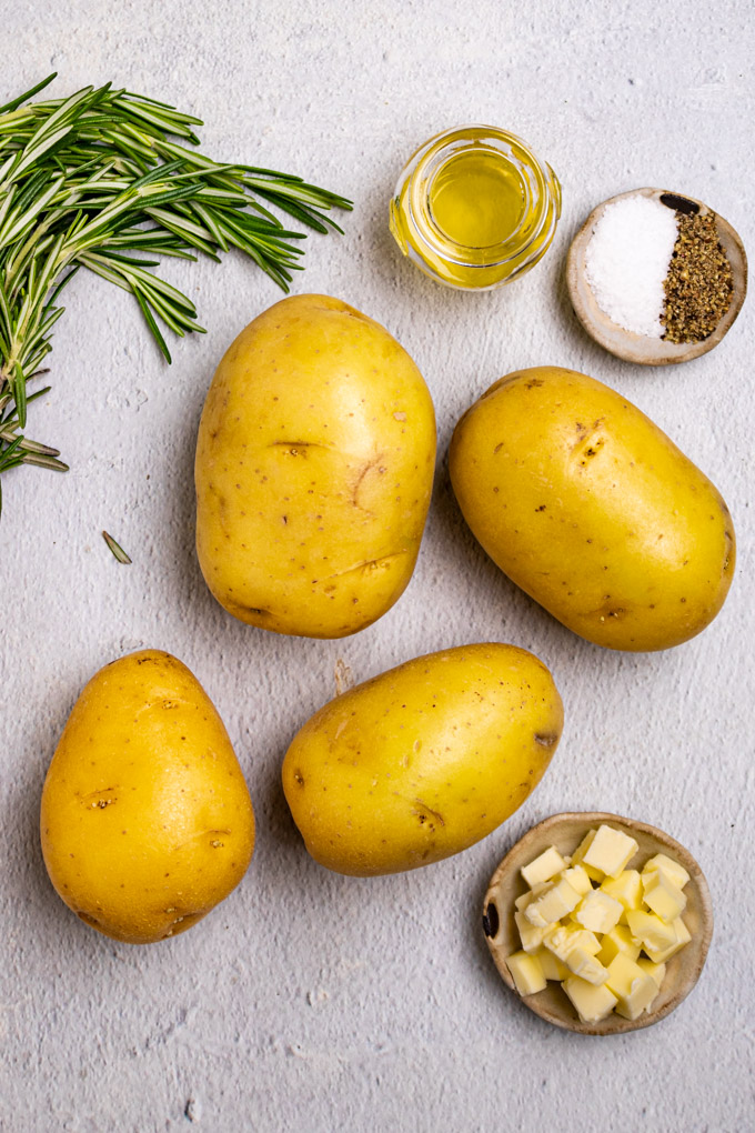 Yukon gold potatoes, rosemary springs, bowl of oil, salt and pepper, and cubes of butter are arranged on gray background.