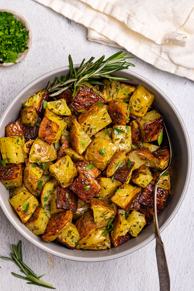 A bowl of roasted potatoes with a spoon sticking in the bowl. A sprig of rosemary is sticking out the side of the bowl.