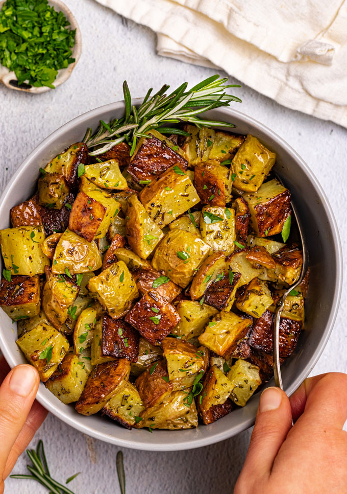 Roasted potatoes are in a gray bowl with two hands holding the bowl. Parsley is sprinkled over the potatoes.