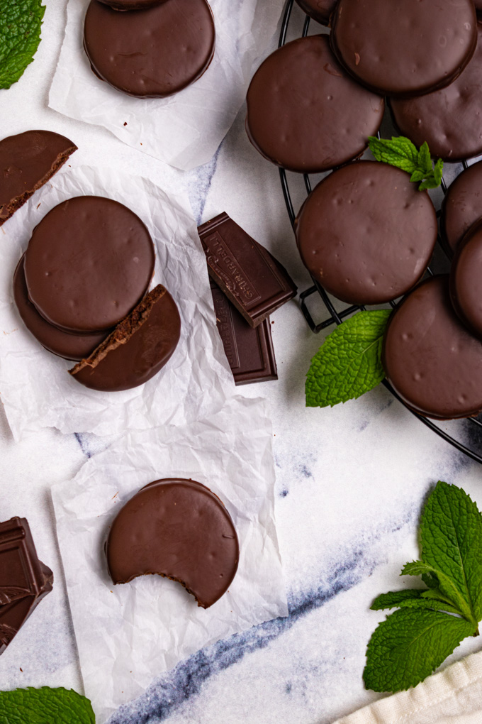 Gluten free thin mints on a blue marble background. One has a bite taken out of it, others are scattered around. There is a round cooling rack off to the corner with more cookies piled on it. Peppermint leaves are also scattered around.