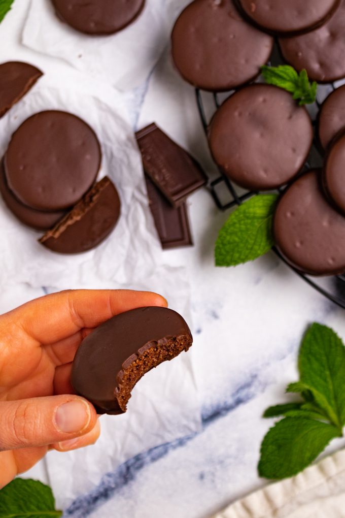 A hand is holding a gluten free thin mint in the air. A bite is taken out of the thin mint. There are extra thin mint cookies scattered around. Some are piled onto a round cooling rack. There are mint leaves scattered around.
