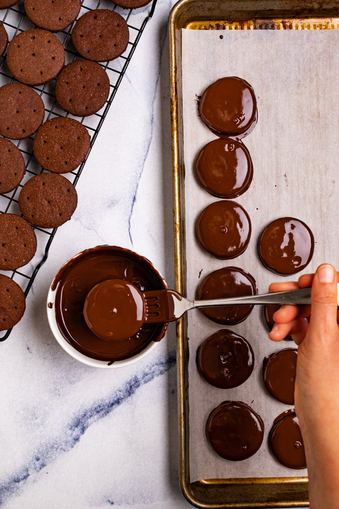 A hand is using a fork to dip a thin mint cookie into a small bowl of chocolate. A baking sheet is to the right with more cookies freshly dipped into chocolate - a cooling rack is to the left with un-dipped cookies.