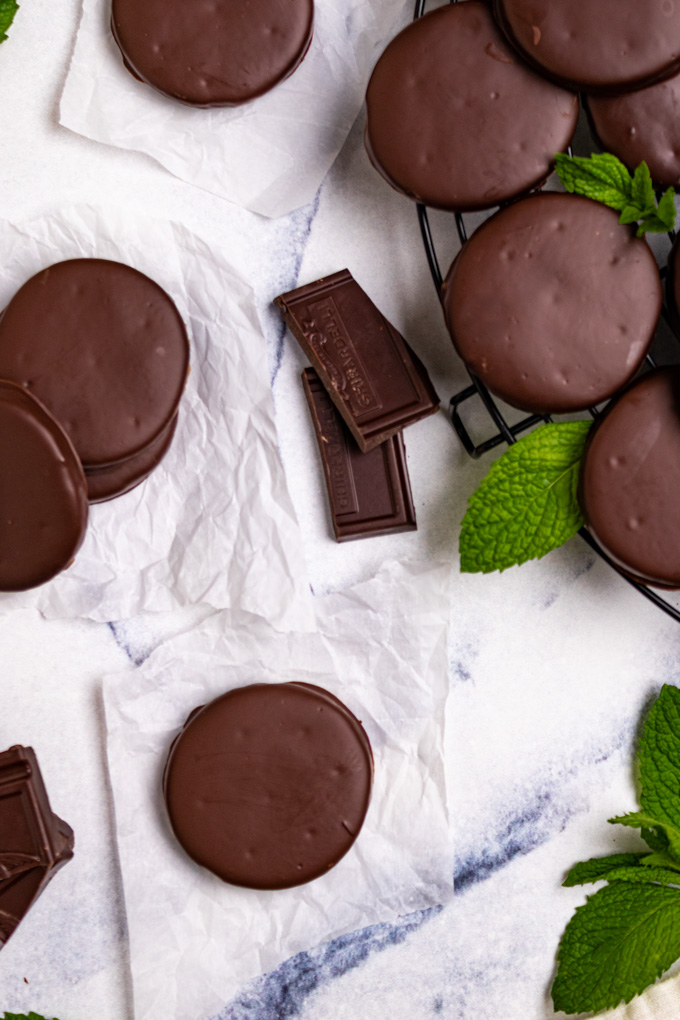 Gluten free thin mints on a blue marble background. Thin mints are strategically placed. There is a round cooling rack off to the corner with more cookies piled on it. Peppermint leaves are also scattered around.