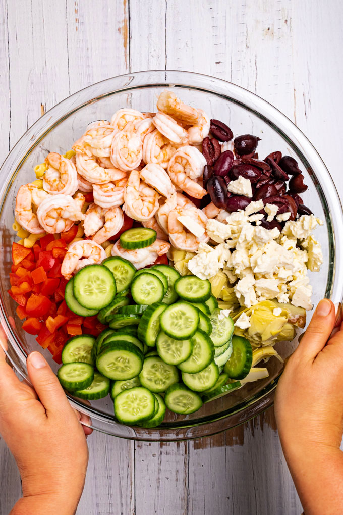A mixing bowl of Greek pasta salad ingredients in groupings. Cucumbers, feta cheese, shrimp, kalamata olives. Hands are holding the bowl.