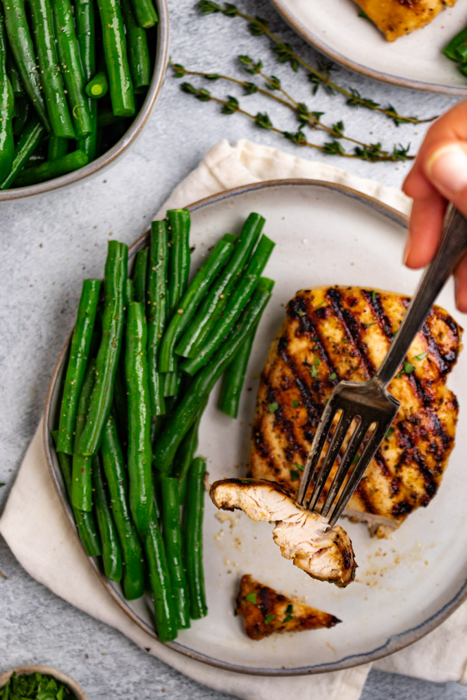 Overhead shot of Italian marinated chicken with a slice of chicken on a fork, being help up close to the camera. Green beans are on the plate, next to the chicken.