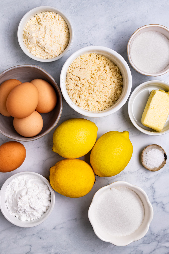 Ingredients for lemon bars in bowls: almond flour, lemon bars, eggs, butter, sugar, tapioca flour, coconut flour.