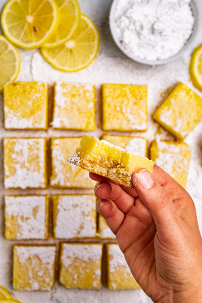 Overhead shot of gluten free lemon bars arranged in a tiled formation. A hand is holding a lemon bar up close to the camera. The lemon bar has a bite taken out of it.