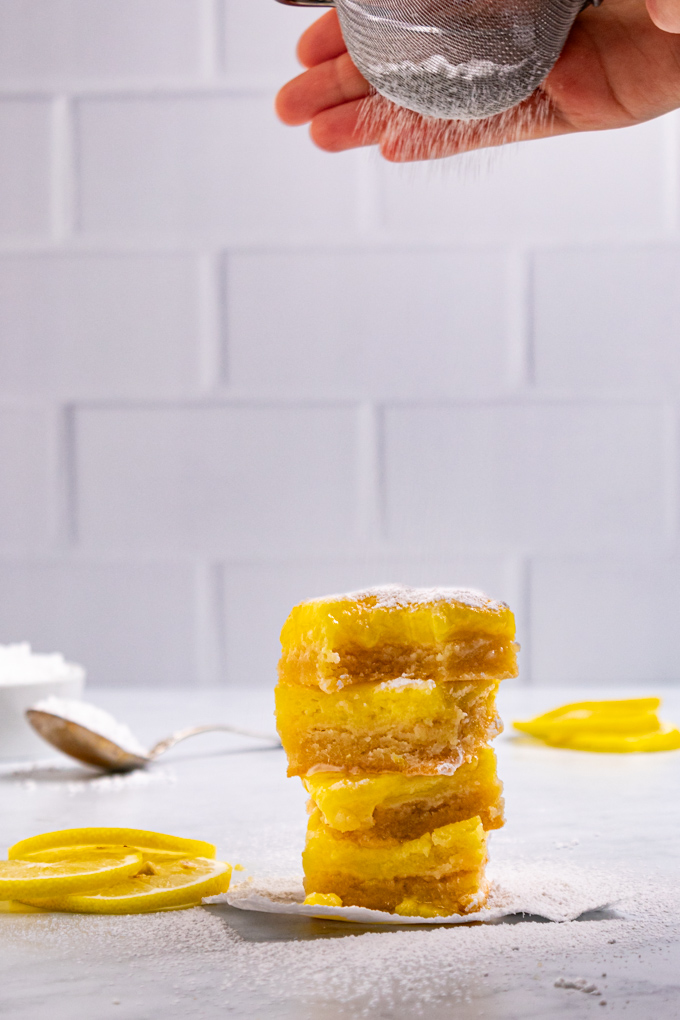 A stack of gluten free lemon bars. The top lemon bar has a bite taken out of it. A hand is holding a fine mesh strainer with powdered sugar over top of the bars, the powdered sugar is being sprinkled on top of the bars.