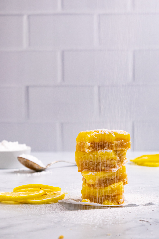 A stack of gluten free lemon bars with a white subway tile background behind it. Powdered sugar is being sprinkled onto the lemon bars. A bowl of powdered sugar is in the background.