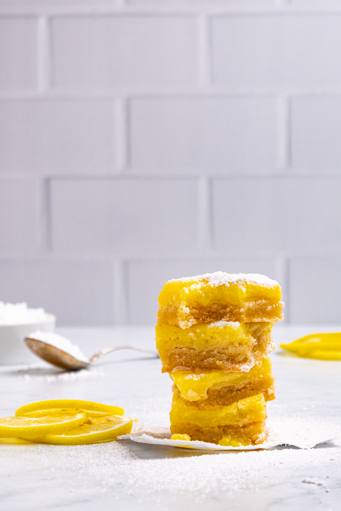 A close up stack of gluten free lemon bars with a bite taken out of the top one. Some powdered sugar is sprinkled on top of the lemon bar. A subway tile backdrop is in the background.