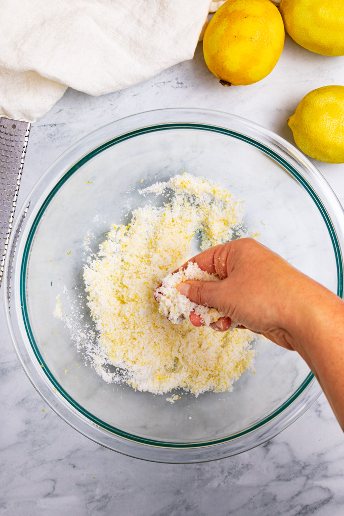A hand is rubbing together lemon sugar in a mixing bowl. A few zested lemons and a white towel, along with a microplane are off to the side.