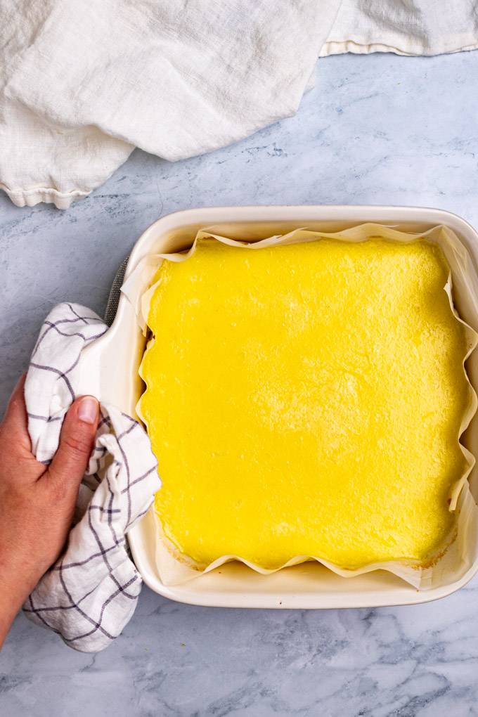 Baked lemon bars are in a white baking dish. A hand is holding the baking dish with a towel.