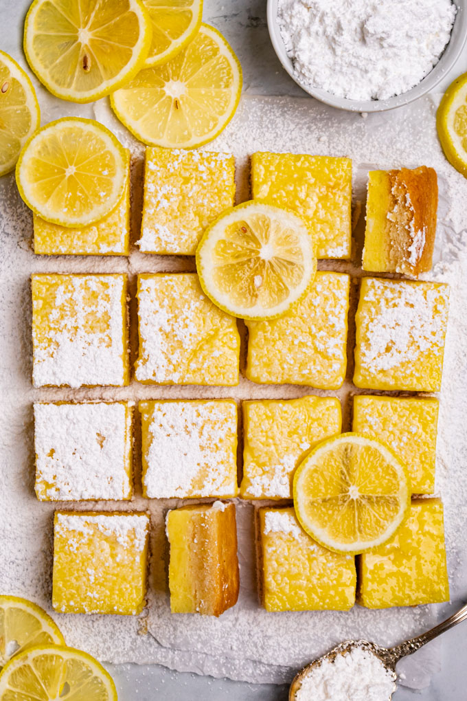 Gluten free lemon bars positioned to make a square pattern. A few of the bars are turned on their side. The bars are dusted with powdered sugar, and have some lemon slices on them. More lemon slices are off to the side, along with a bowl of powdered sugar, and a spoonful of powdered sugar.