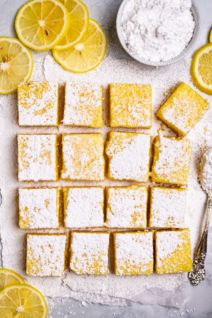 gluten free lemon bars cut into squares with powdered sugar dusted over top. Lemon slices are scattered off to the side, a bowl of powdered sugar is off to the side as well.