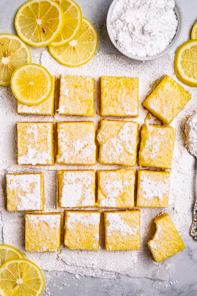 Overhead shot of gluten free lemon bars on a marble counter. The lemon bars are arranged in a square formation with powdered sugar sprinkled on top. There are a few lemon bars askew, one of them has a bite taken out of them. Lemon slices are scattered about. There is a bowl of powdered sugar off to the side.
