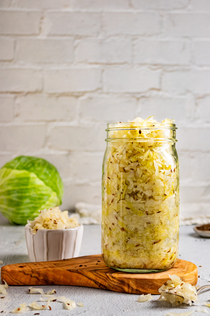 Straight on shot of homemade sauerkraut in a large mason jar. The jar is on a wooden board, there is a small fork with more sauerkraut laying next to the jar, a small bowl of sauerkraut is off the the side, and a head of green cabbage is in the background. The backdrop is white brick.