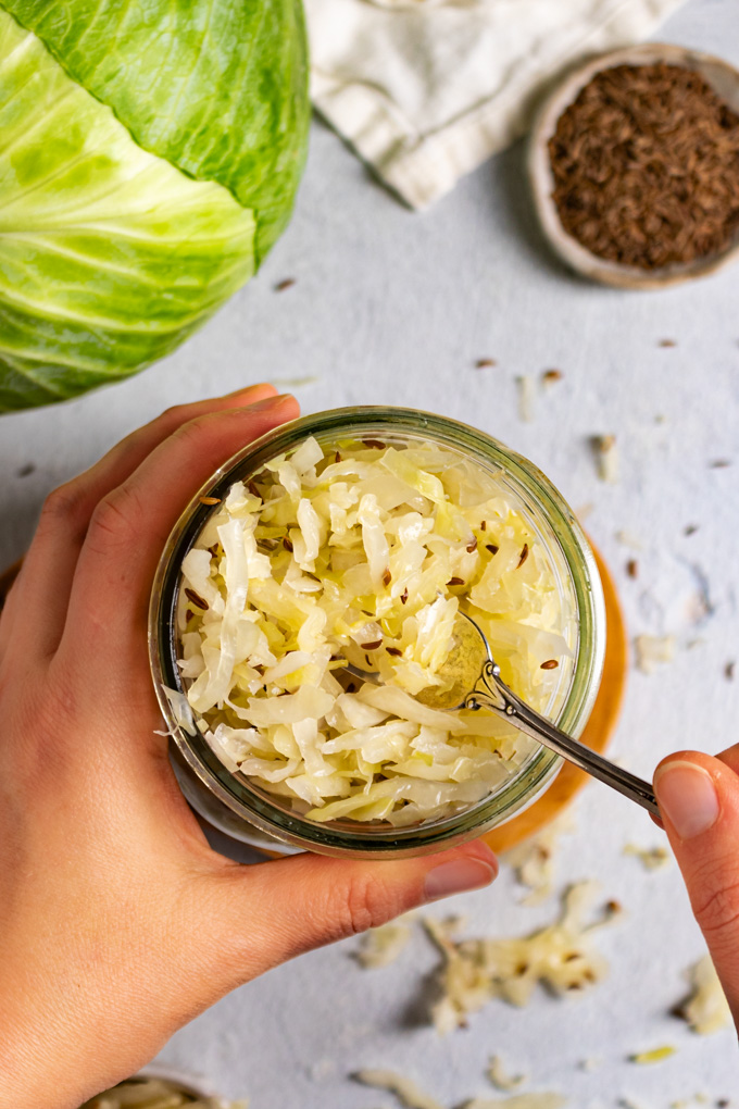 Overhead shot of sauerkraut in a mason jar with hands holding the mason jar, and a fork dipping into the sauerkraut.