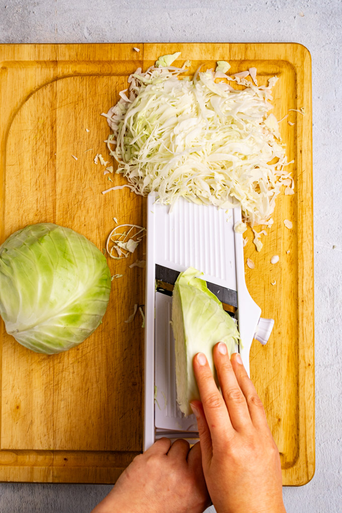 Slicing a head of cabbage on a mandolin. A hand is holding a piece of cabbage, and shredded cabbage is piled at the end of the mandolin.
