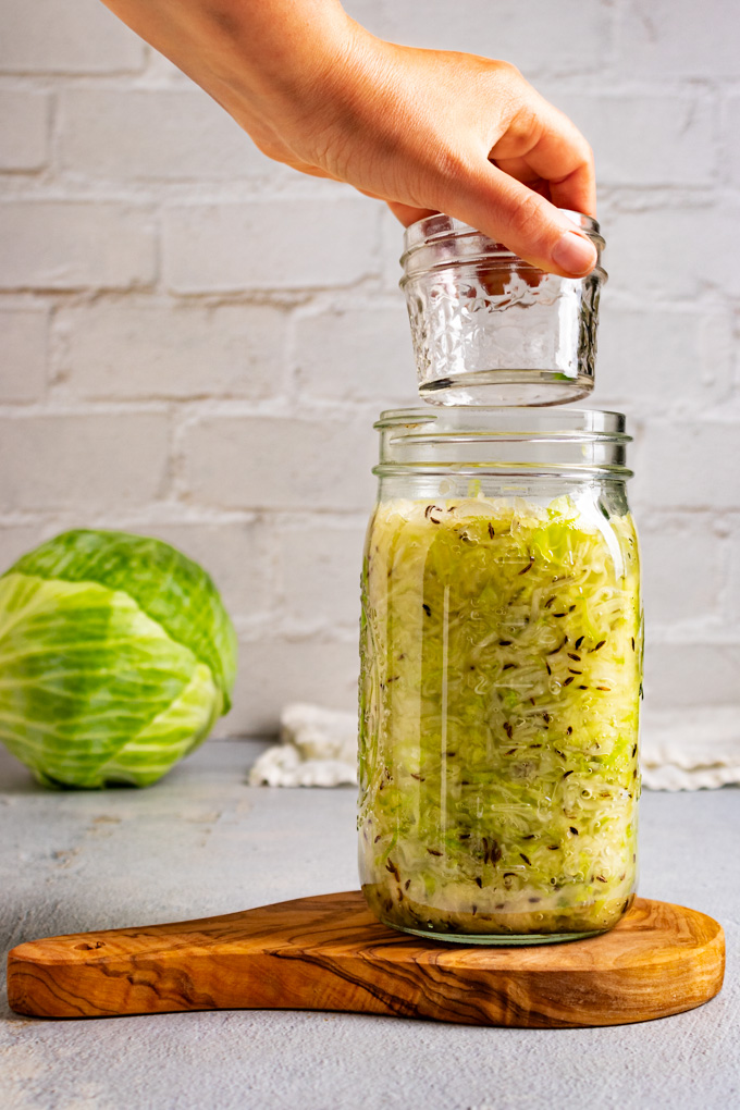 Straight on shot of a small mason jar being added to a large mason jar full of shredded cabbage, to weigh down the cabbage to be fermented into sauerkraut.