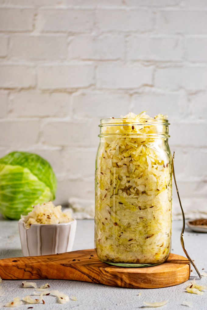How to make sauerkraut - Straight on shot of sauerkraut in a mason jar. There is a fork leaning on the mason jar, a small bowl of sauerkraut, and a head of cabbage in the background. There is white brick as the backdrop.