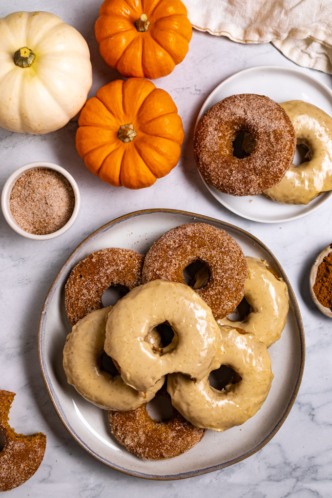 Overhead photo of baked pumpkin donuts. The donuts are stacked on a plate, the top one has a bite taken out of it. The donuts have a either a sugar coating on them, or a maple glaze. Another small plate of donuts is in the upper right corner. There are mini pumpkins in the upper left corner. In the lower left corner there is a sugared pumpkin donut with a bite taken out of it.