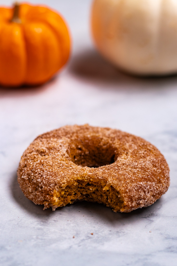 A close up angled shot of a single baked pumpkin donut that is covered in a sugar mixture. A bite is taken out of the donut. There are mini pumpkins in the background that are out of focus.