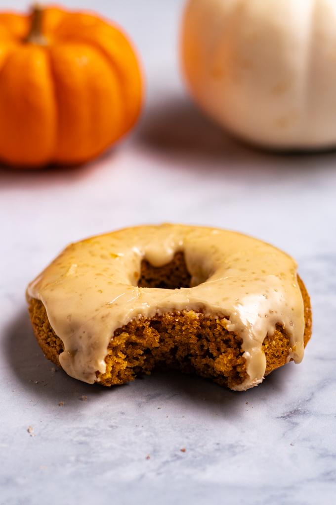 A close up angled shot of a single baked pumpkin donut that is covered in a maple glaze. A bite is taken out of the donut. There is glaze dripping down the sides of the donut. There are mini pumpkins in the background that are out of focus.