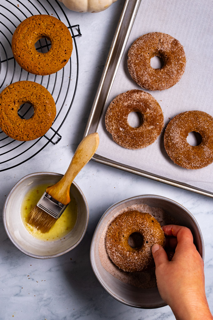Overhead shot of pumpkin donuts being covered in sugar. A hand is holding a donut in the sugar mixture, an other bowl has melted butter, and a pastry brush in it. The upper left corner has a cooling rack with plain donuts on it, and a baking sheet has donuts covered in sugar on it. 