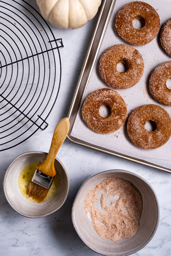 Pumpkin donuts covered in sugar on a baking sheet. A bowl of melted butter with a pastry is in the left bottom corner. Another bowl of sugar and pumpkin spice in the bottom right hand corner.