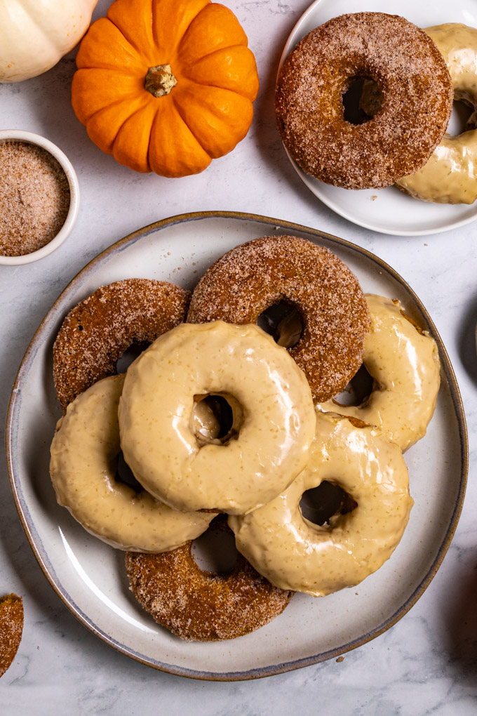 Overhead photo of baked pumpkin donuts. The donuts are stacked on a plate. Some are covered in a sugar coating, others are coated in a maple glaze. There is another small plate of donuts in the upper right hand corner. There are mini pumpkins in the upper left corner.
