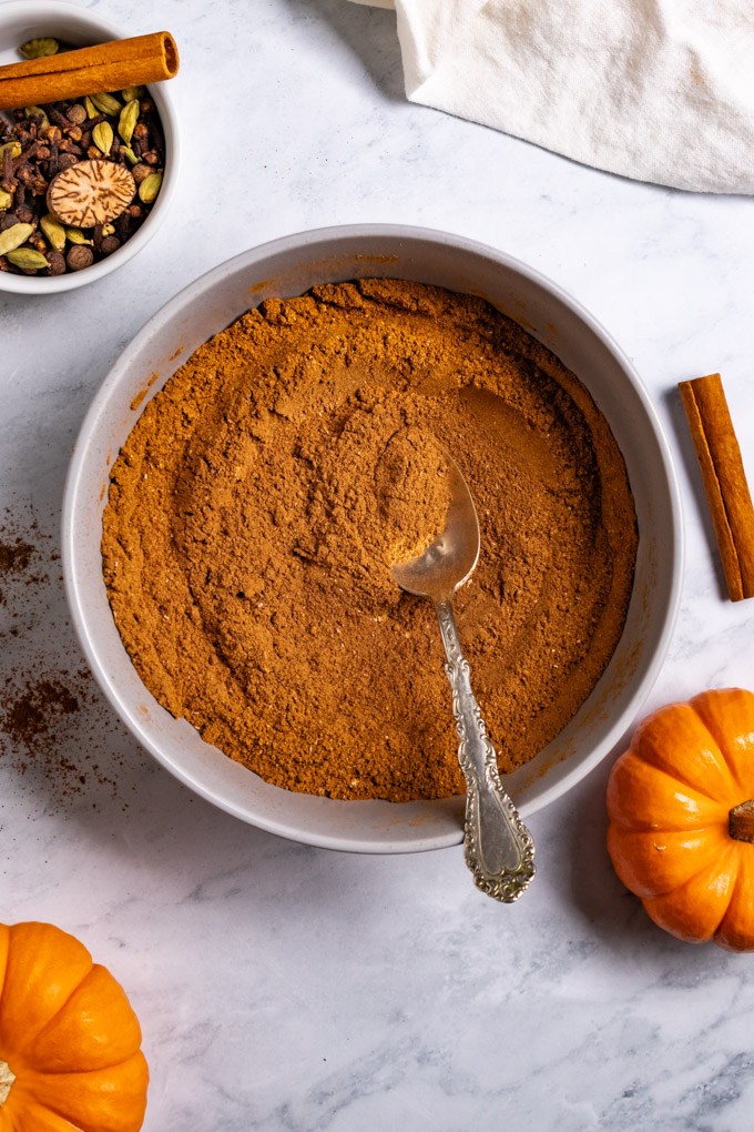 Overhead photo of homemade pumpkin pie spice in a gray bowl with a spoon in it. There are mini pumpkins off to the side, along with a small bowl of whole spices, and a cinnamon stick.