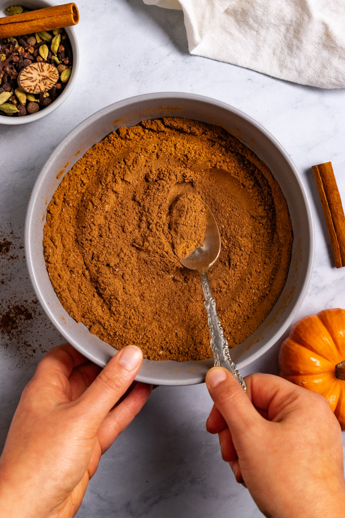 Pumpkin pie spice is in a gray bowl, with a spoon in it. Hands are holding the bowl, and the spoon. The whole spices, are in the corner of the photo, along with a cinnamon stick. A mini pumpkin is in the bottom right corner.