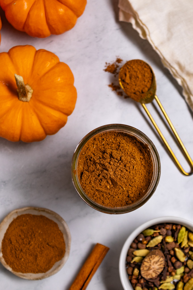 Overhead photo of homemade pumpkin pie spice in a jar. A measuring spoon has more pumpkin pie spice in it, off to the side, along with whole spices in a bowl, and a cinnamon stick. Two mini pumpkins are in the corner of the photo.