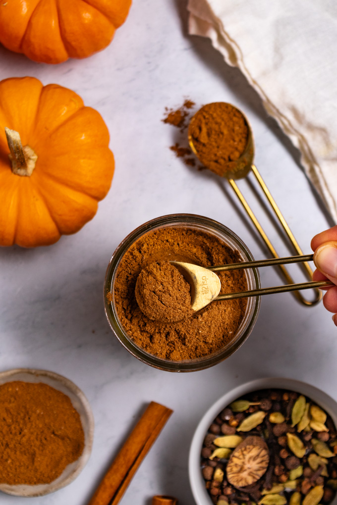 Overhead photo of homemde pumpkin pie spice in a jar. A hand is holding a measuring spoon full of pumpkin pie spice over the jar. There is a cinnamon stick in the bottom left corner, mini pumpkins in the top left corner, and a bowl of whole spices in the bottom right corner. 
