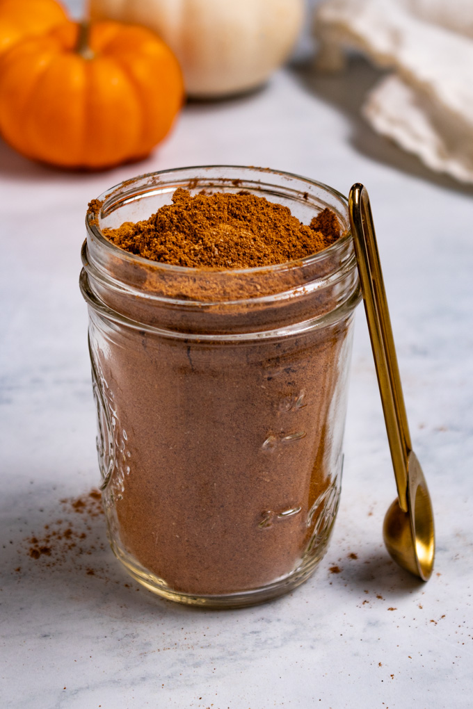 Angled photo of homemade pumpkin pie spice in a jar. A measuring spoon is balancing against the mason jar. Mini orange pumpkins, and a white mini pumpkin are in the background.