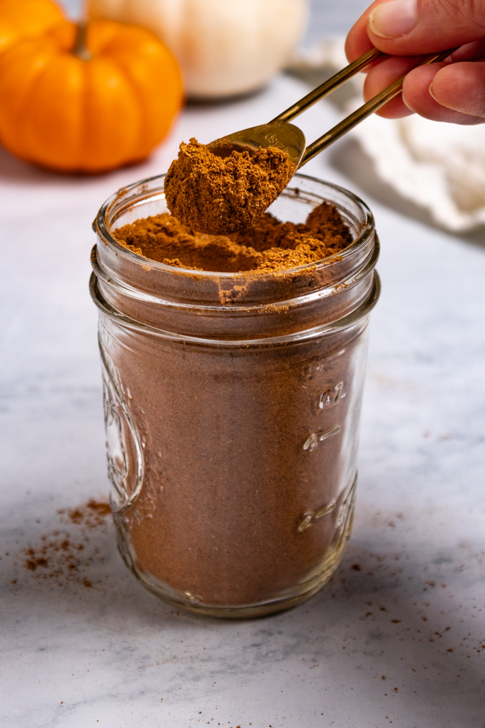 Homemade pumpkin pie spice is in a jar. A hand is holding a measuring spoon in the air, with some pumpkin pie spice in it. Mini pumpkins are in the background.